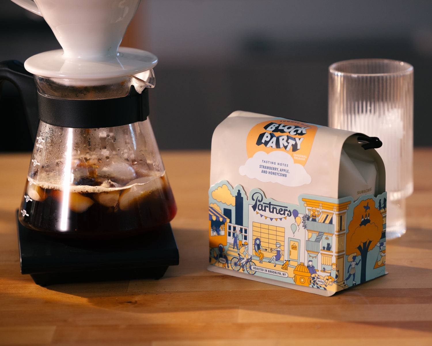 Three-quarter view of Block Party coffee bag showing colorful illustrated packaging and tasting notes next to an iced flash-brew carafe on a wooden counter