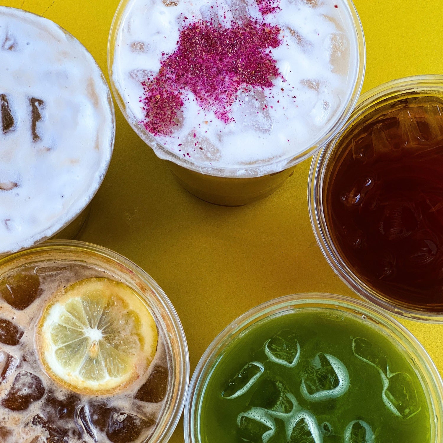 Five iced coffee drinks on a yellow table: lemon-topped drink, green matcha, dark iced coffee, and two foam-topped drinks