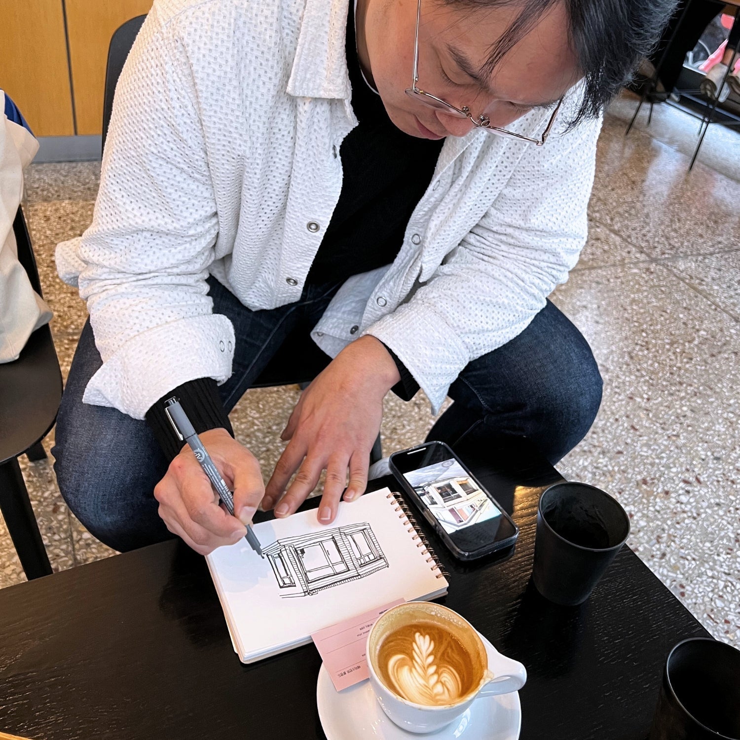 Artist sketching a cafe storefront in a spiral notebook at a coffee shop table, phone and latte with heart-shaped latte art nearby