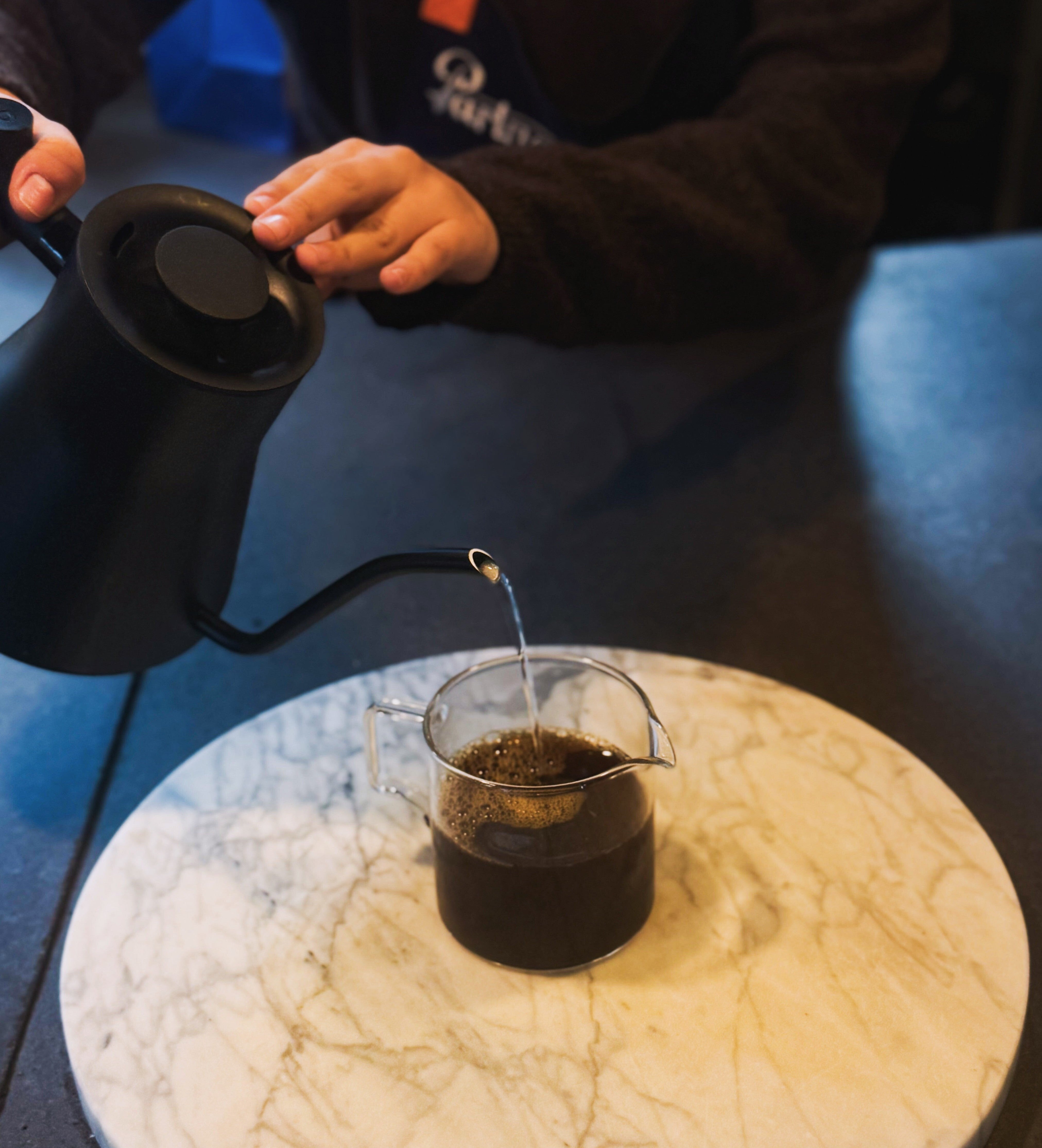 Hands pouring hot water from a black gooseneck kettle into a glass pitcher of brewed coffee on a round marble board