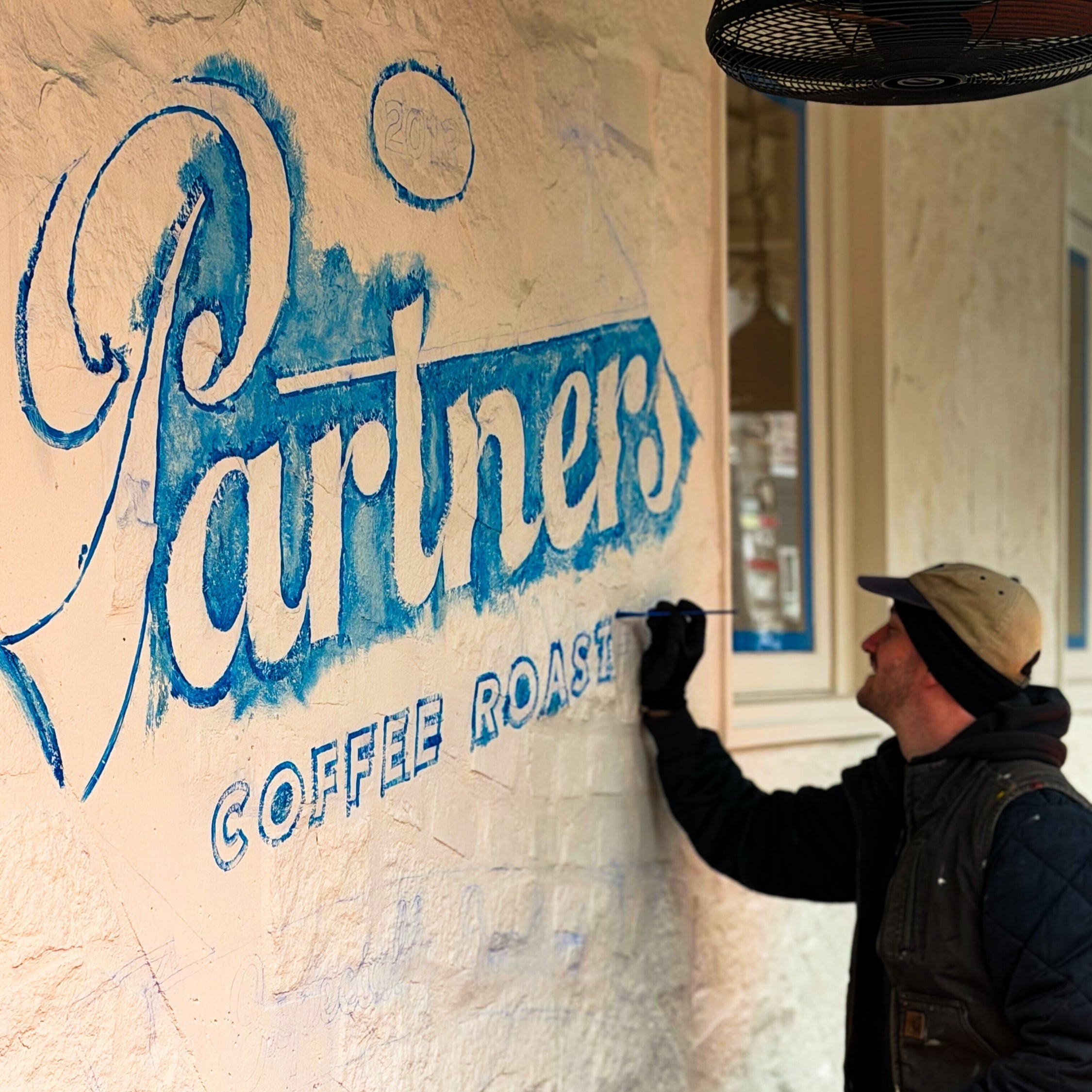 Artist painting ‘Partners Coffee Roasters’ mural on a textured cafe exterior wall