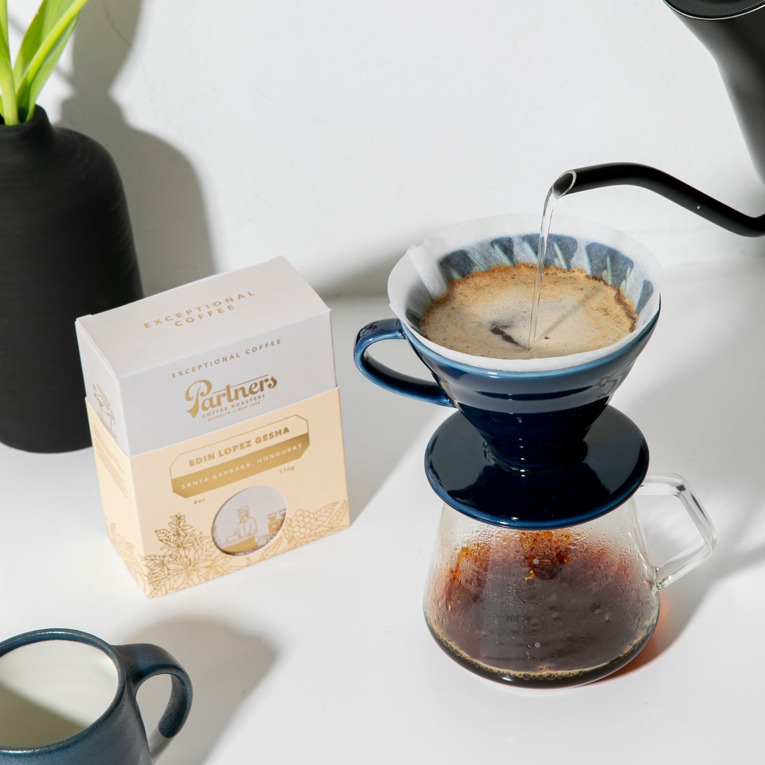 Box of Partners Coffee Edin Lopez Gesha beside a blue V60 pour over brewing into a glass carafe on a white tabletop