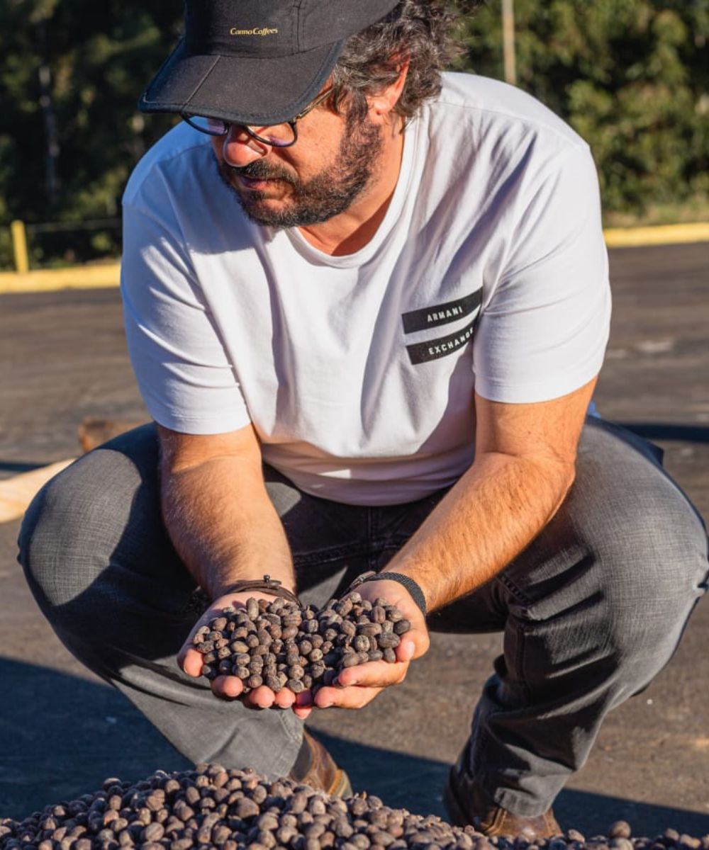 Man crouching outdoors inspecting handfuls of coffee beans over a pile