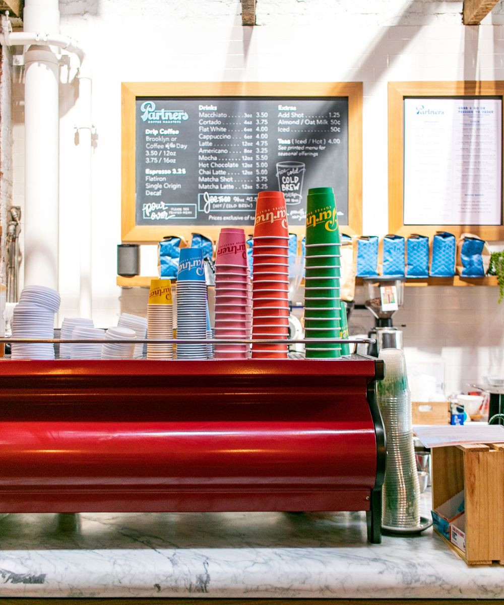 Espresso machine and stacked colorful takeaway cups on a cafe counter with a chalkboard menu in the background