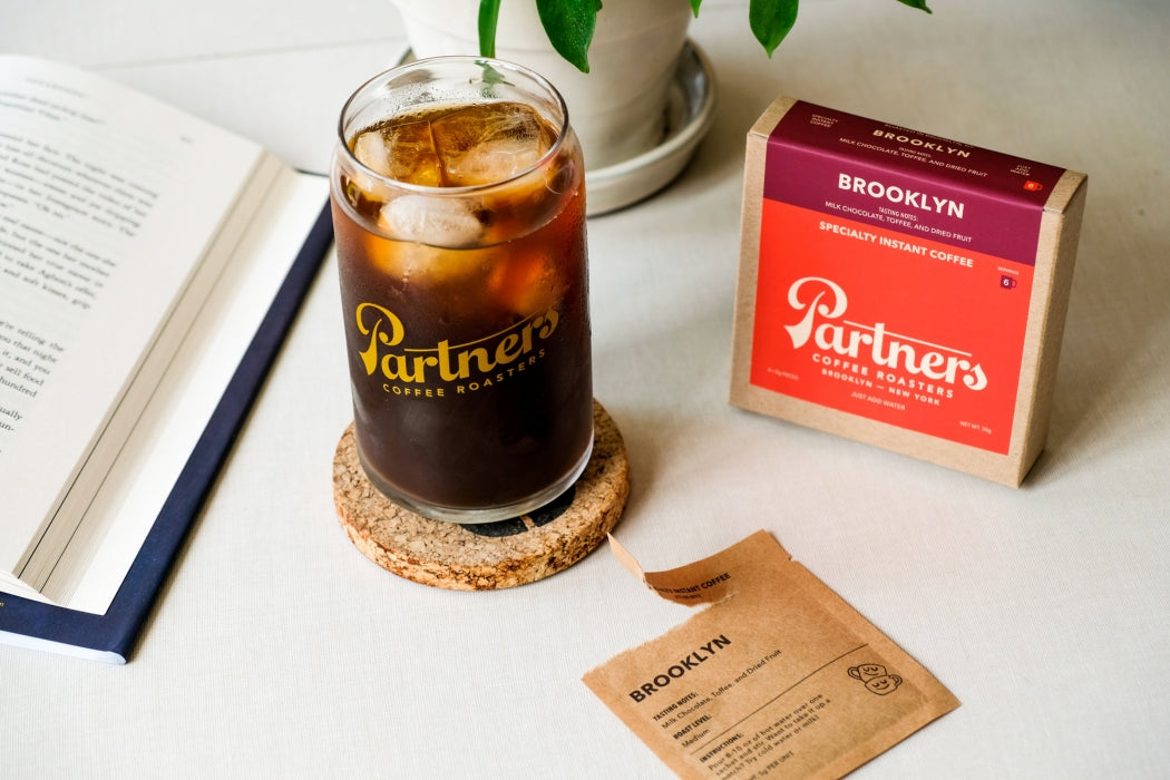 Angled view of Ghost Town Decaf instant coffee box and iced glass with Partners logo and open sachet on light tabletop