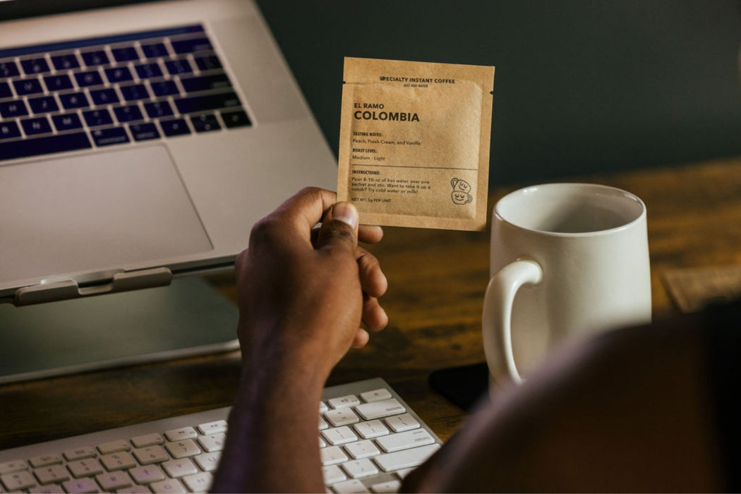 Hand holding a single-serve specialty instant coffee sachet beside a laptop keyboard and white mug on a wooden desk