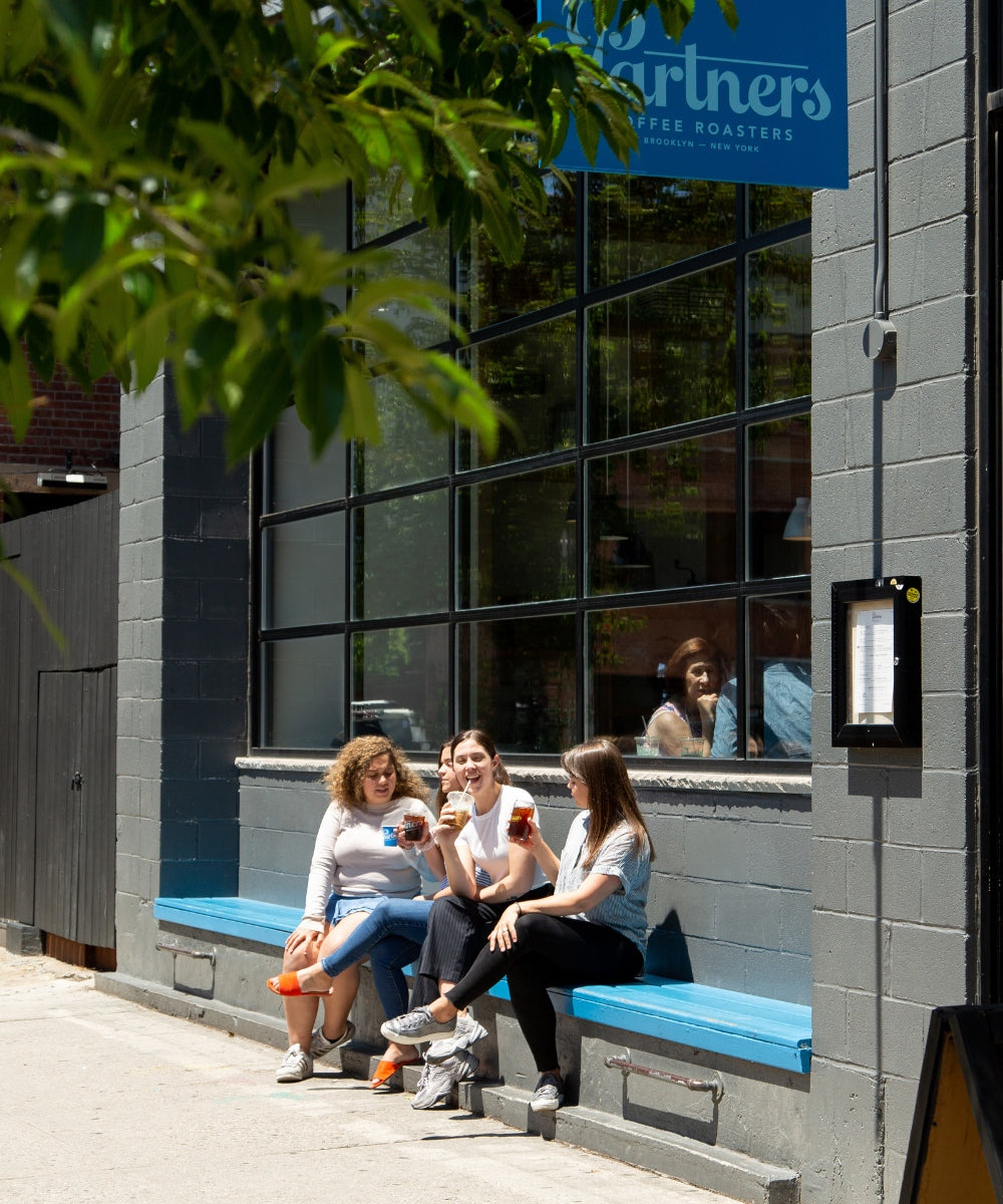 Three people sitting on a blue bench outside Partners Coffee’s Williamsburg roastery, smiling and drinking takeaway coffee on a sunny day