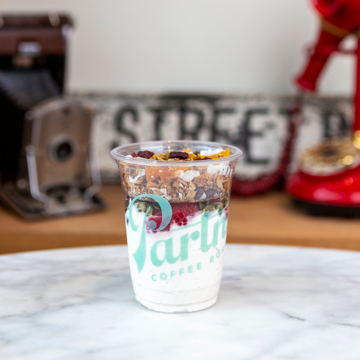 Close-up of parfait cup with layered yogurt, granola, and berries on a marble counter with blurred coffee equipment in background