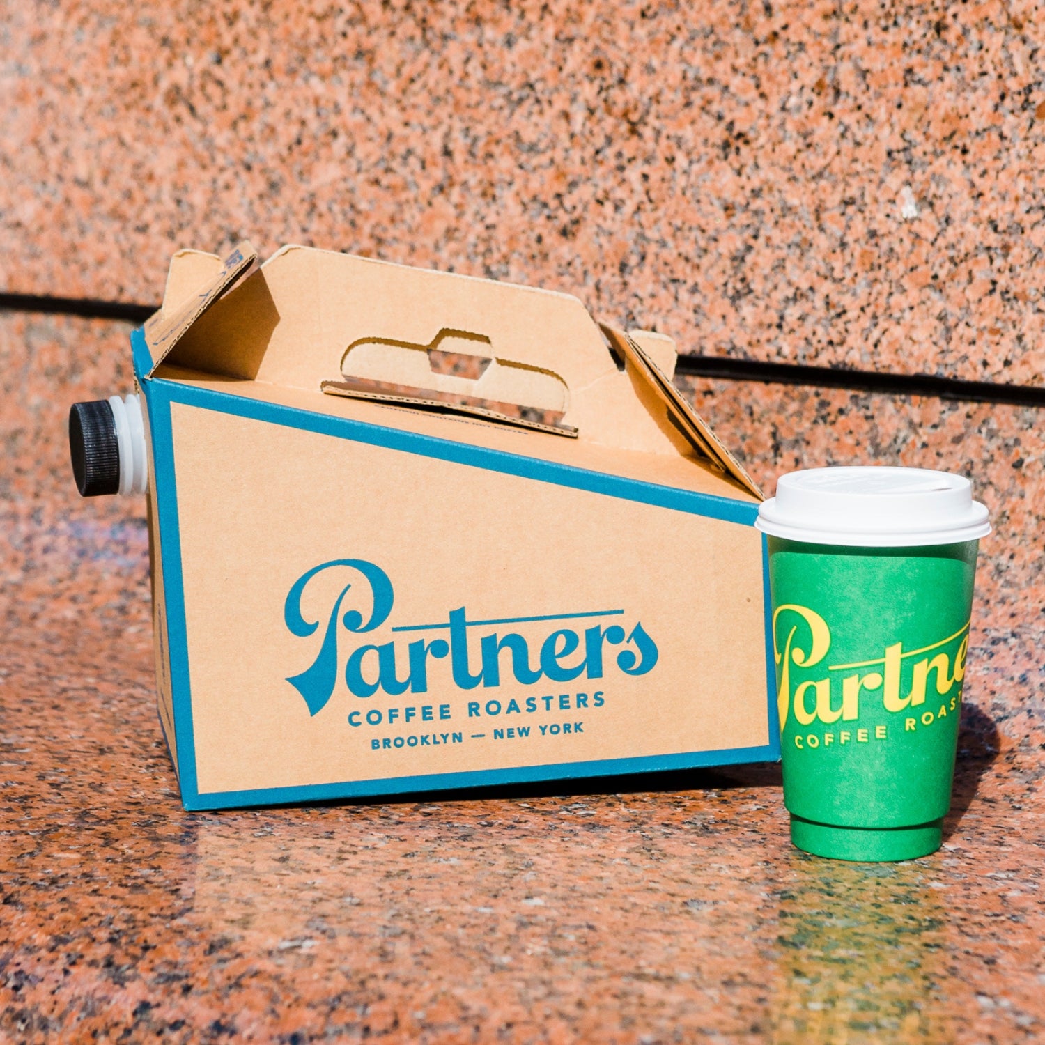 Three-quarter view of Partners catering coffee box with built-in spout and branded green takeaway cup on a stone surface