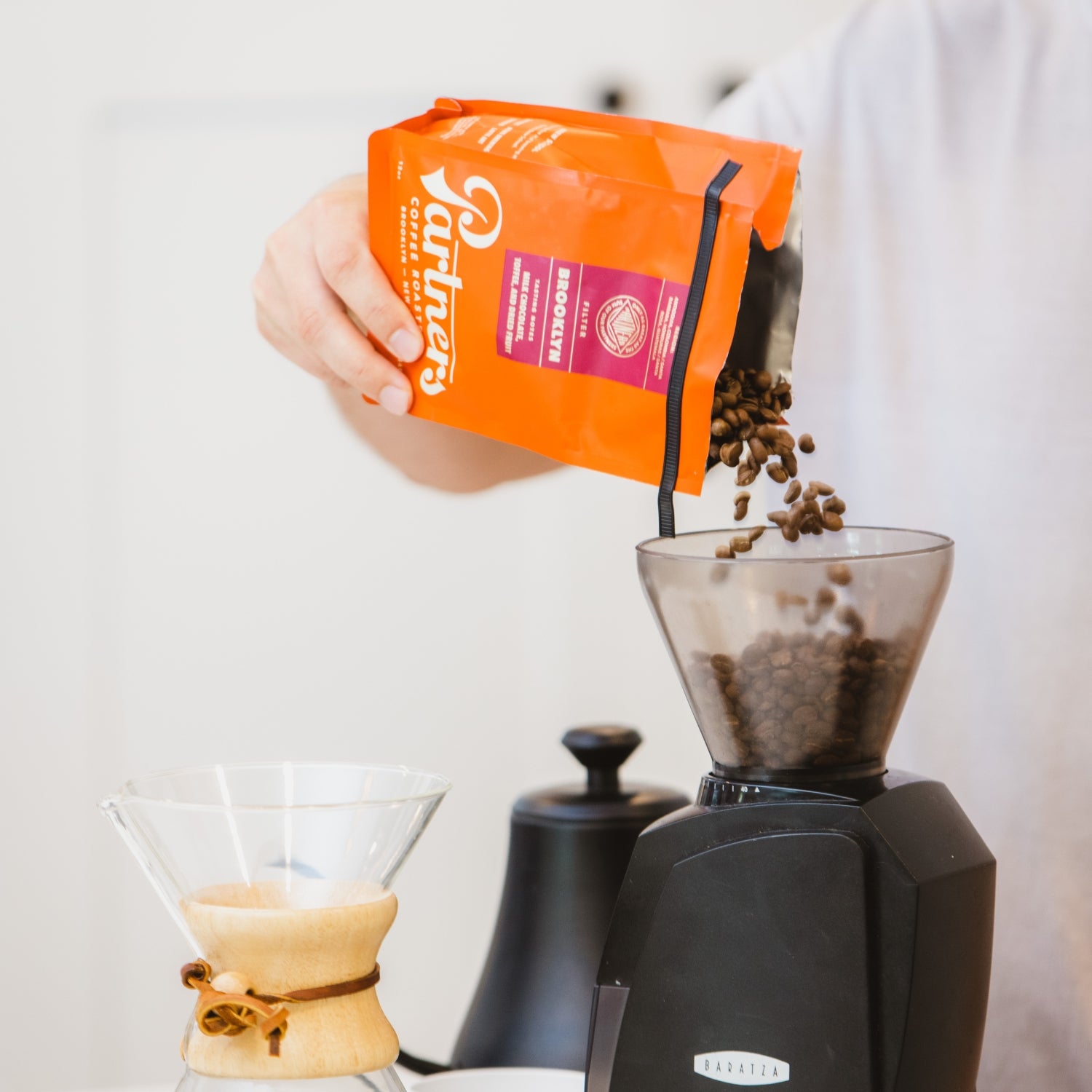 Person pouring Partners Coffee orange bag of roasted beans into a grinder next to a Chemex and kettle in a bright kitchen