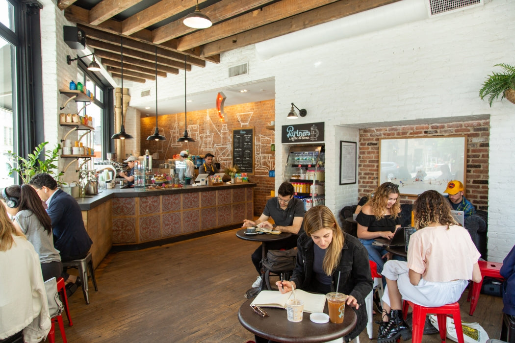 People talking and working at tables in a sunlit coffee shop interior with exposed wooden beams, brick wall, and service counter
