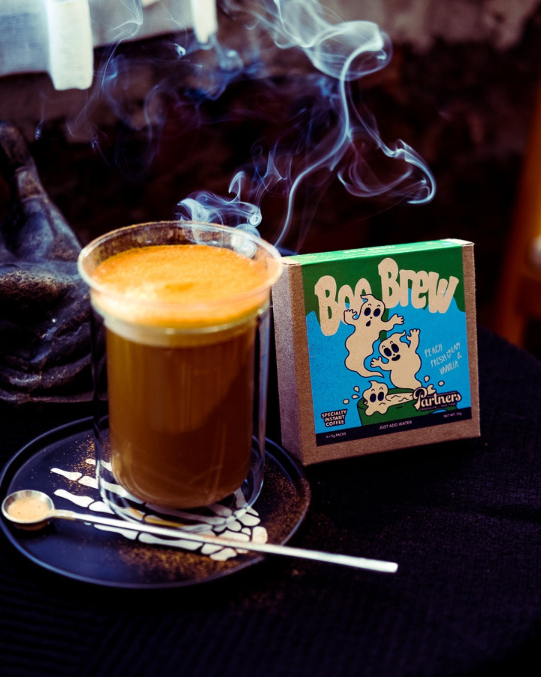 Front view of Boo Brew instant coffee box beside a steaming glass of coffee with frothy crema on a black saucer