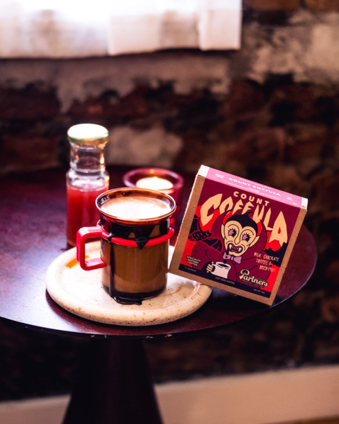 Angled view of Count Coffula instant coffee box beside a steaming mug and lit candle on a table with exposed brick