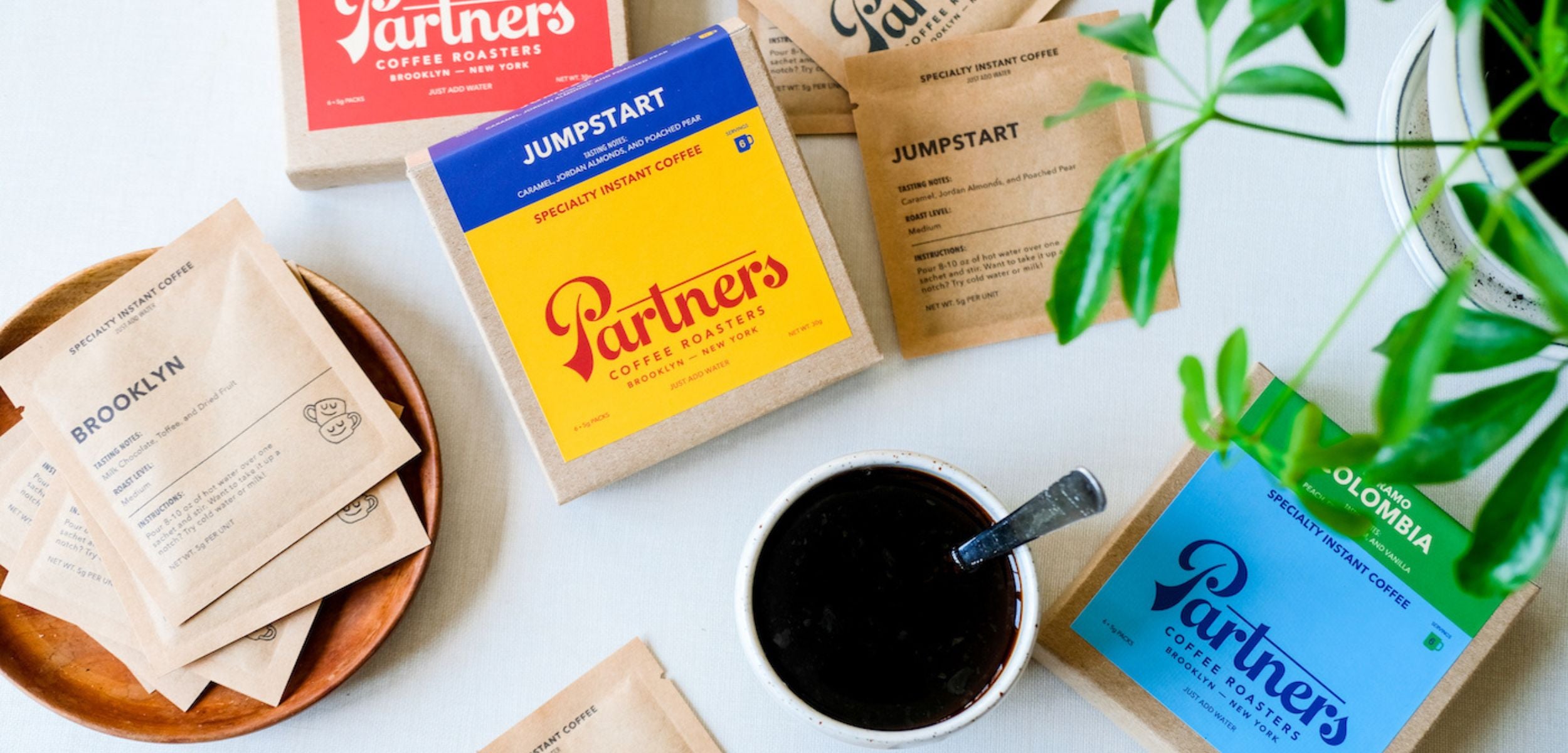 Flat lay of three Partners Coffee specialty instant coffee boxes and single-serve sachets with a brewed cup on a white surface