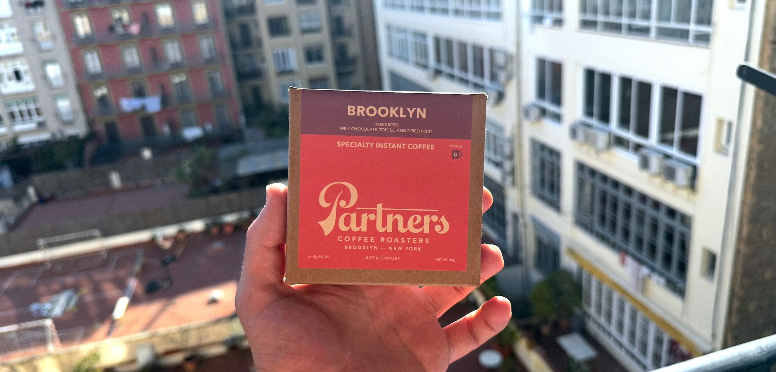 Hand holding a box of Partners specialty instant decaf coffee on a balcony with apartment buildings in the background