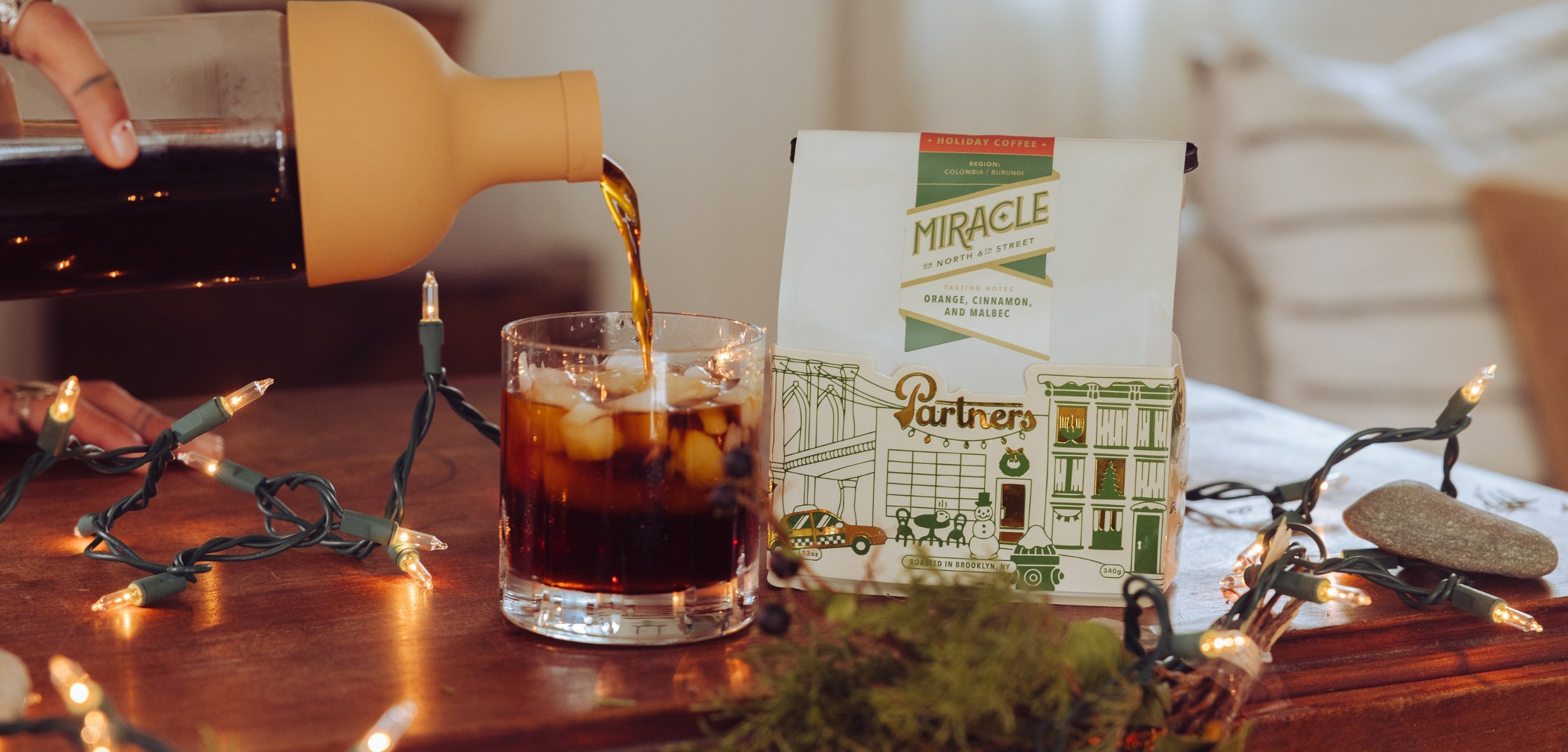 Hand pouring coffee over ice into a glass beside a Partners Coffee Miracle holiday bag and string lights on a wooden table