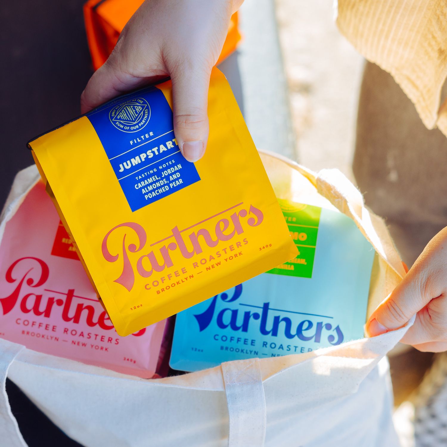 Hands placing a yellow Partners coffee bag labeled Jumpstart into a tote with two other colorful Partners coffee bags