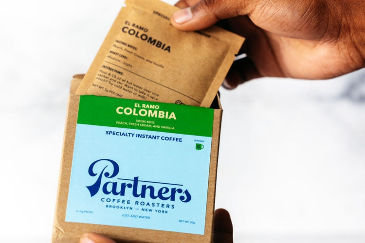 Close-up of two El Ramo Colombia instant coffee sachets with Partners Coffee logo held in hands against a white background