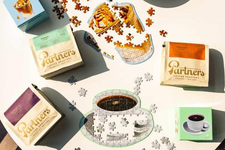 Overhead of four Partners Coffee bags arranged around a partially assembled jigsaw puzzle depicting a coffee cup