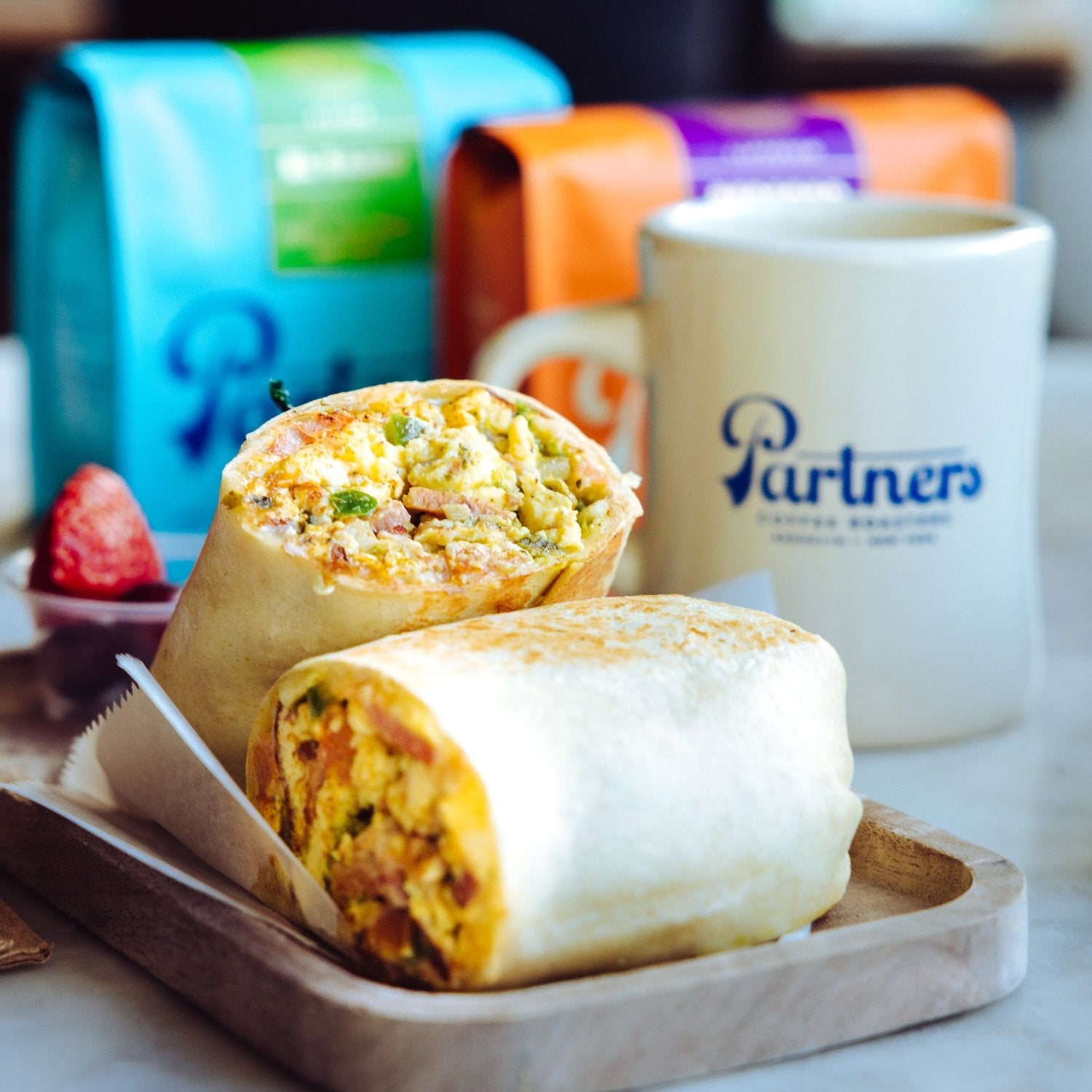Close-up of burrito cut in half showing egg, veggies, and cheese with a Partners coffee mug and bags in background