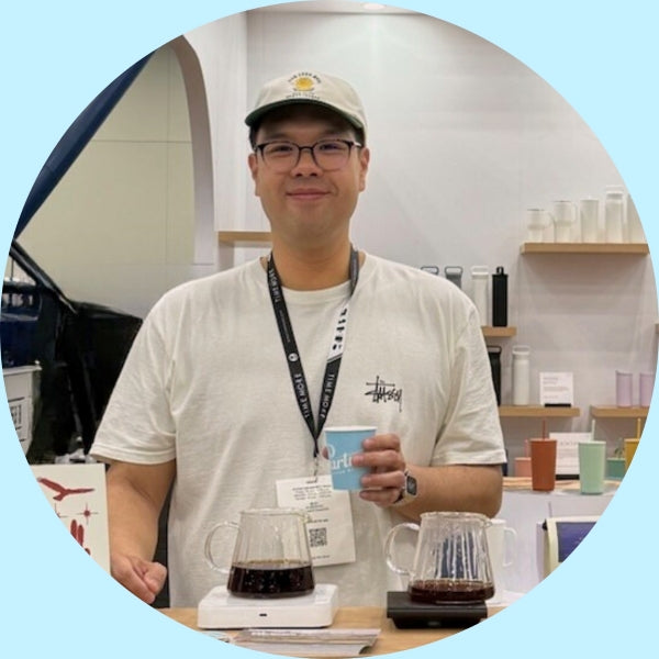 Staff member standing behind a table with two pour-over coffee carafes, holding a blue cup in a bright booth with shelves of colorful cups behind him