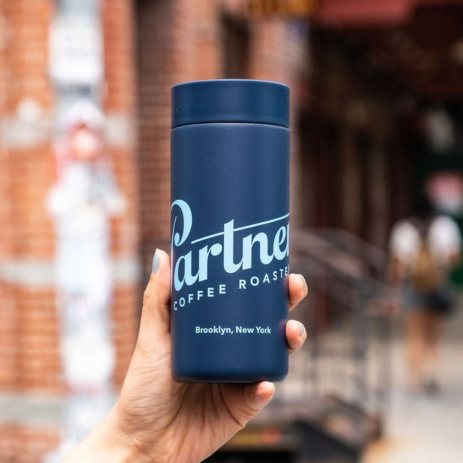 Handheld front view of Partners x MiiR 360 Traveler blue 16oz travel mug showing Partners logo and 360-degree sip lid with blurred city background