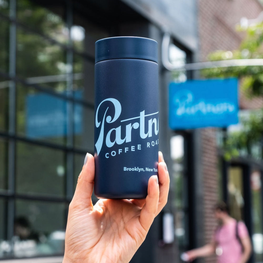 Front view of Partners x MiiR 360 Traveler 16oz blue showing logo and 360-degree lid, handheld outside cafe