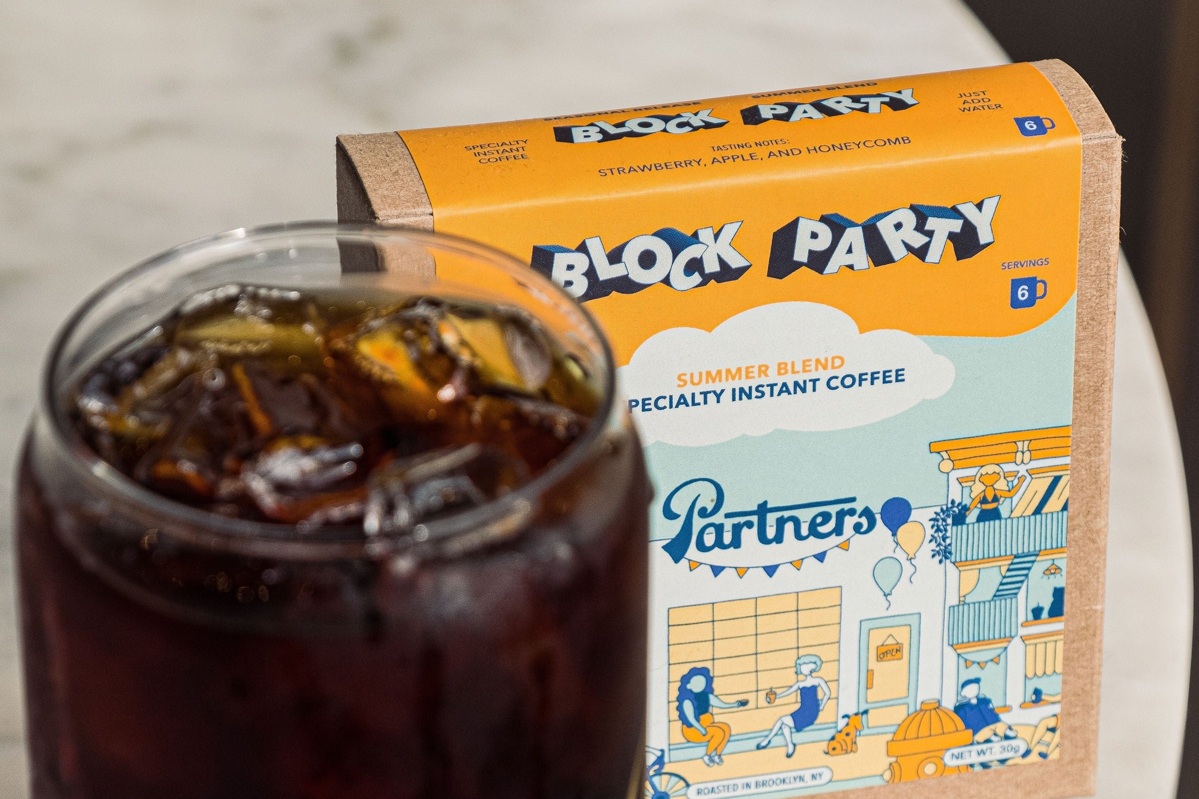 Front view of Block Party Summer Blend instant coffee box with Partners logo and a glass of iced coffee on a marble surface