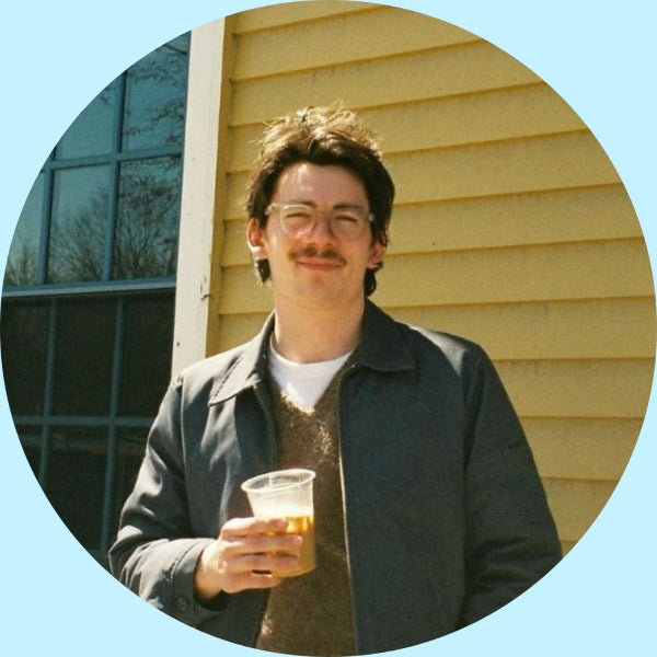 Drew, Partners Coffee digital marketing and graphic designer, smiling and holding a cup of iced coffee outside a yellow clapboard building