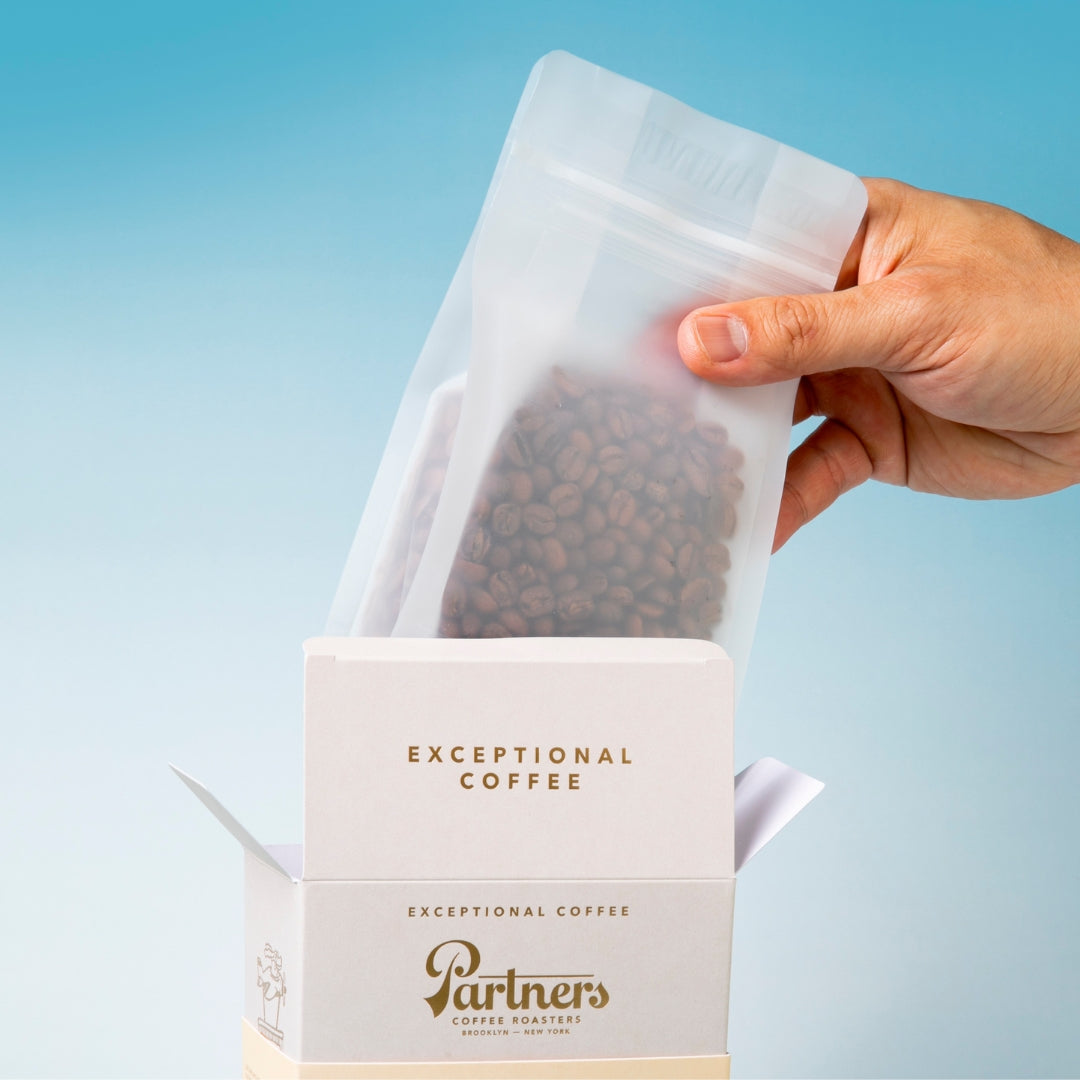 Hand inserting resealable pouch of Edin Lopez Gesha coffee beans into open Partners Coffee box on pale blue background