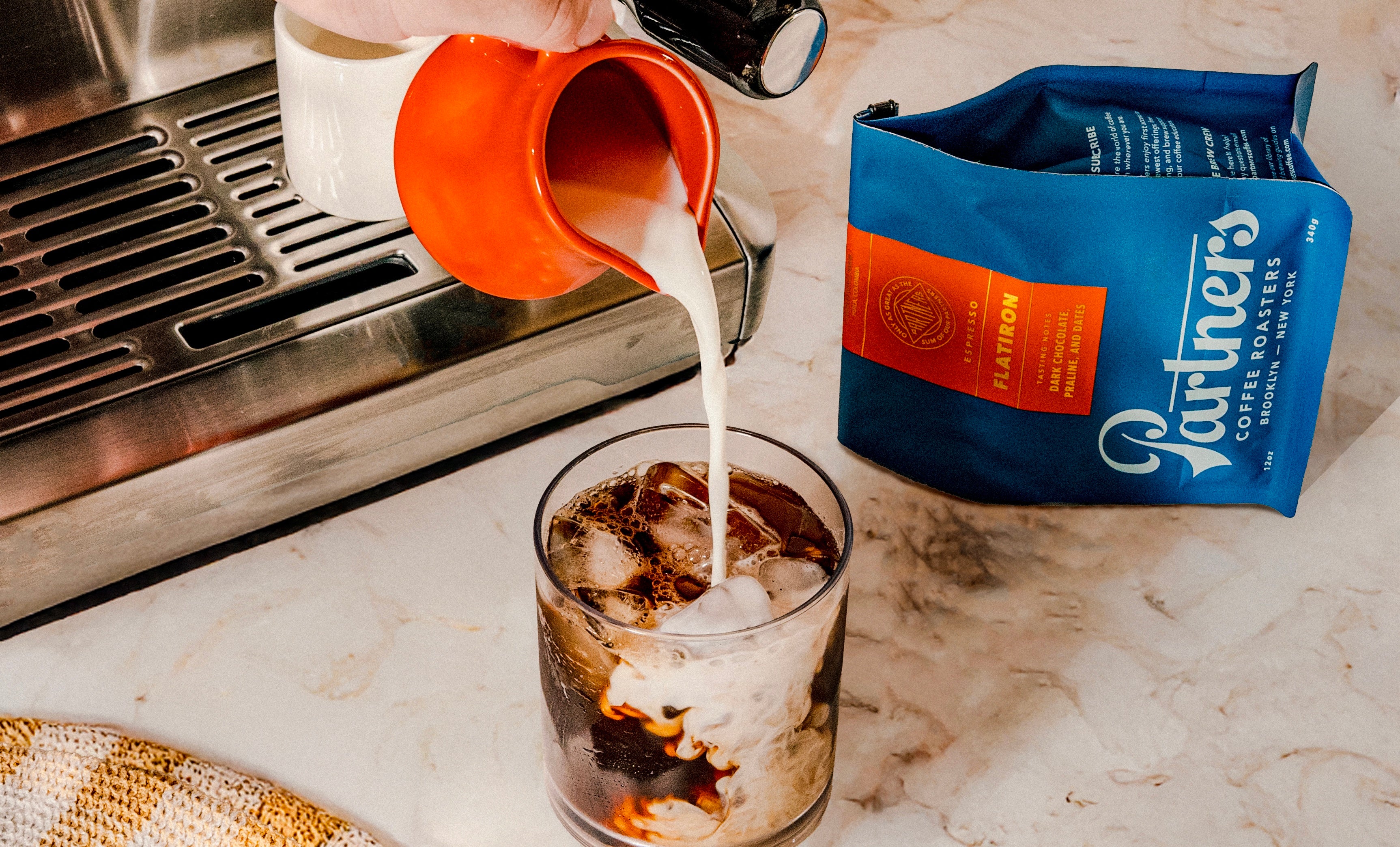 Hand pouring milk into a glass of iced coffee beside a blue Partners Flatiron coffee bag on a marble counter