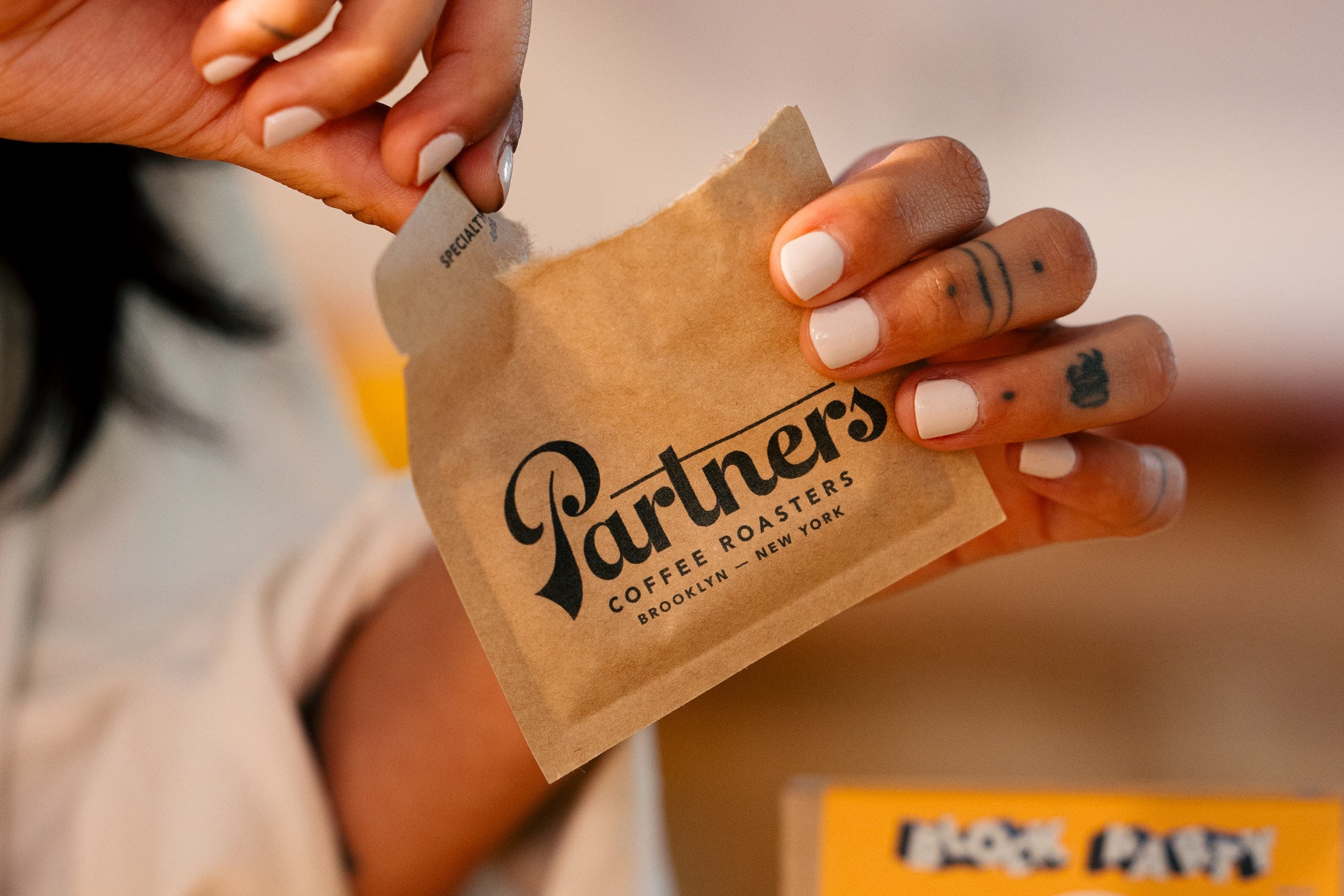 Hands tearing open a Partners Coffee Roasters instant coffee sachet with a blurred mug and Block Party box in the background