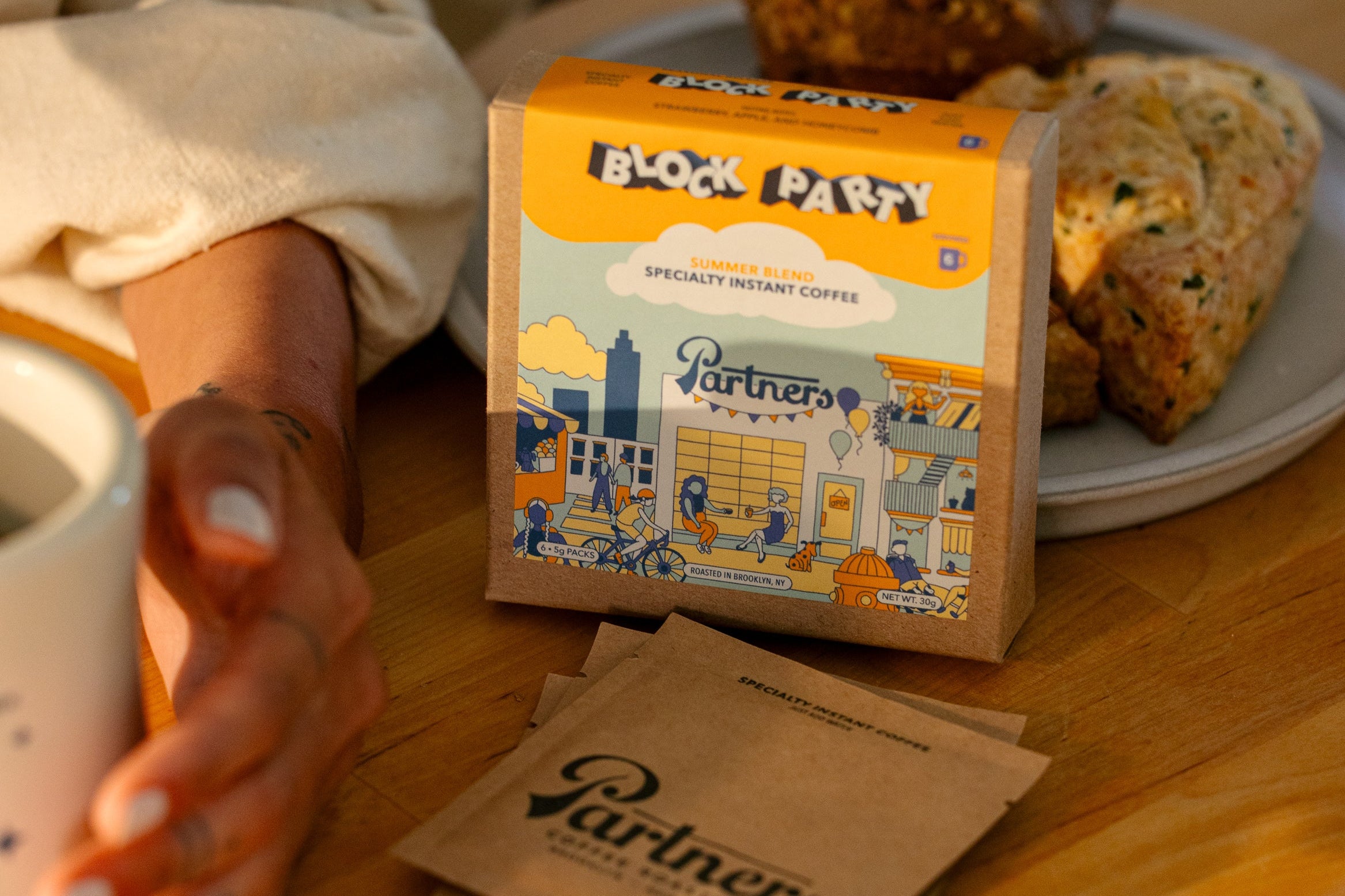 Box of Block Party specialty instant coffee sachets with Partners Coffee branding beside a mug and scone on a wooden table in warm sunlight