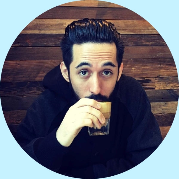 Team member sipping a small espresso from a clear glass while seated against a wood-paneled background