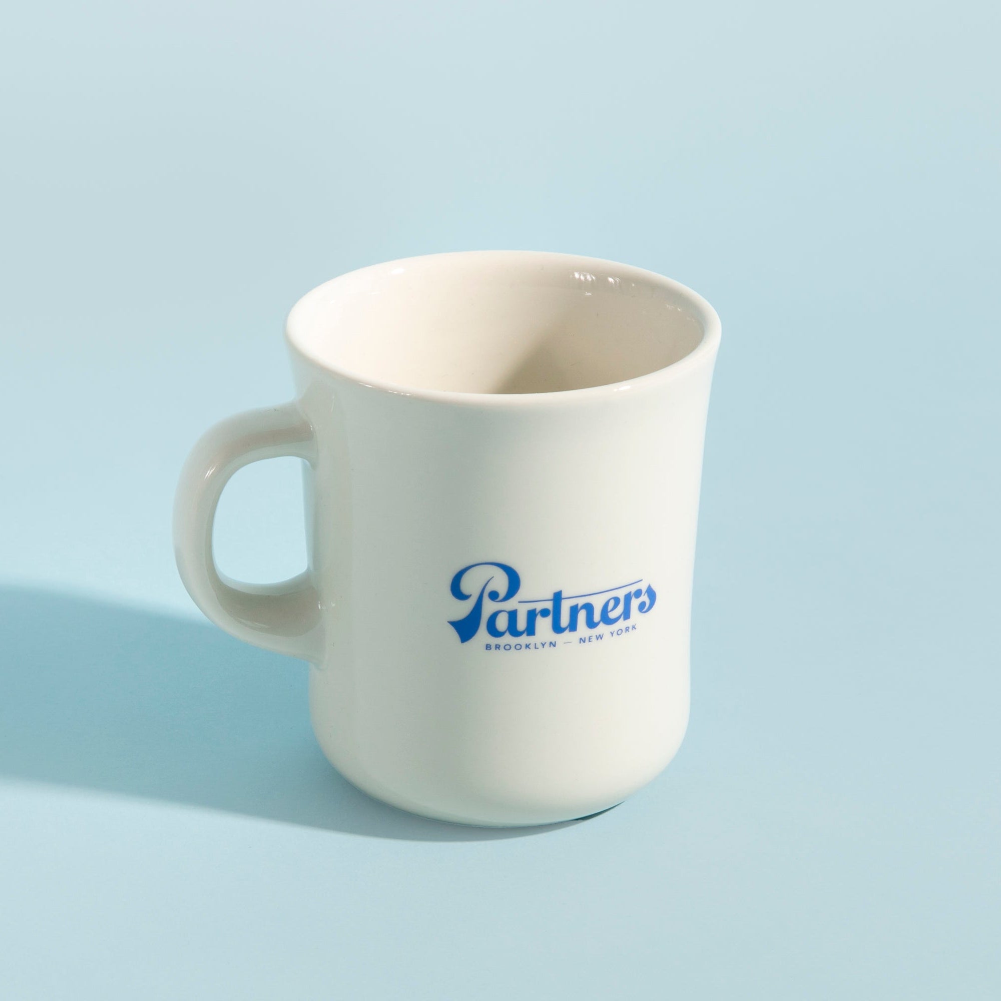 Angled front view of Partners x Kinto Diner mug showing blue Partners logo on white porcelain with pale blue background