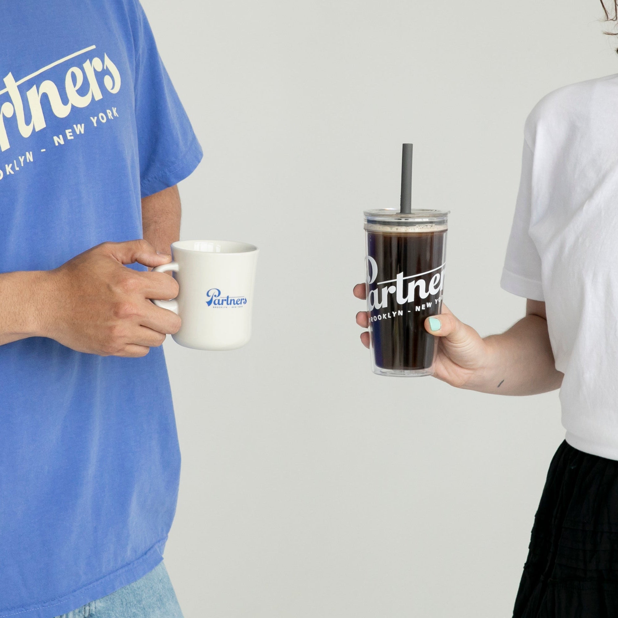 Two people holding drinks: a white Kinto Diner porcelain mug in one hand and a clear Partners tumbler with a straw against a neutral background