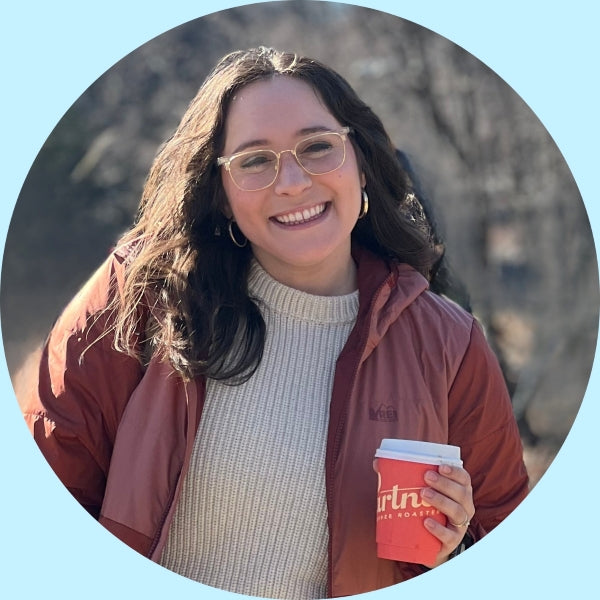 Woman smiling outdoors holding a red Partners cup, wearing glasses, cream sweater and rust-colored jacket