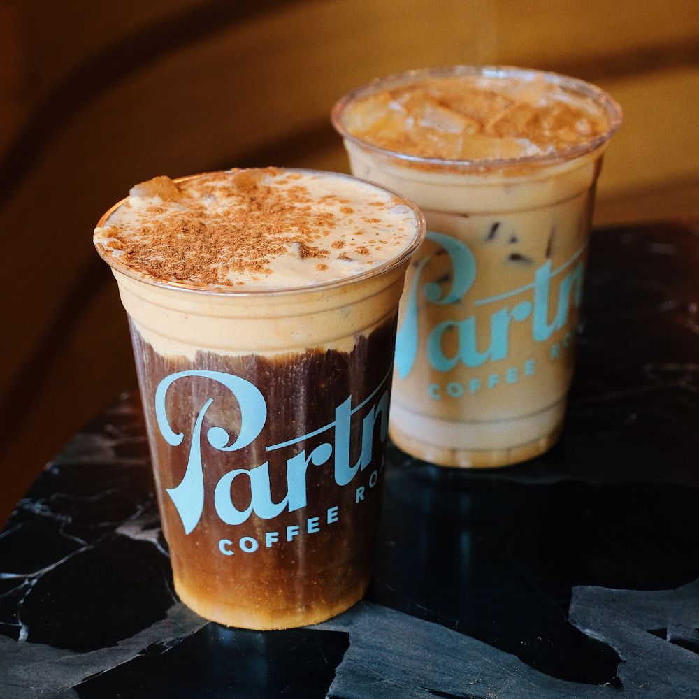 Three-quarter view of an Iced Pumpkin Pie Latte behind a Pumpkin Cold Foam Cold Brew, showing layered dark cold brew topped with pumpkin foam and a cinnamon sprinkle on a dark marble table