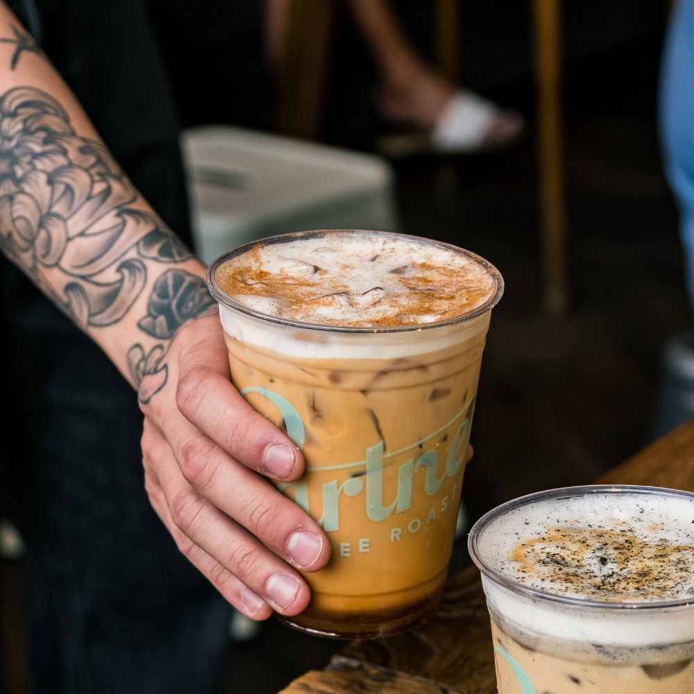 Tattooed hand lifting a Partners Coffee plastic cup of iced latte with foamy, cinnamon-dusted top at a cafe counter