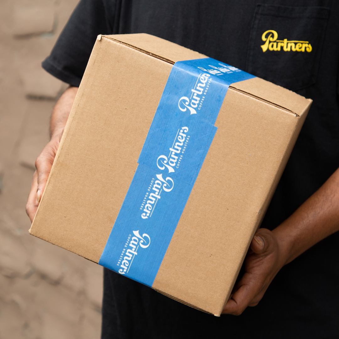 Person holding a cardboard delivery box sealed with blue tape reading Partners, wearing a black Partners shirt