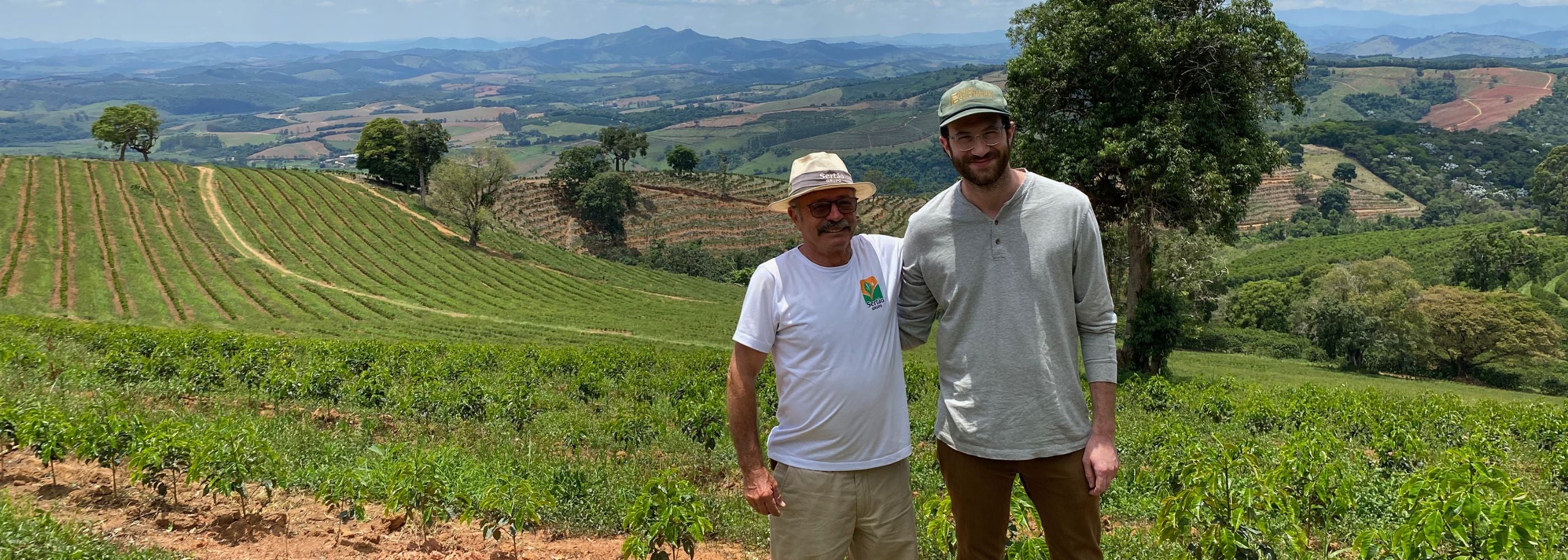 Two people standing on a coffee farm in Brazil among rows of coffee plants with rolling hills in the background