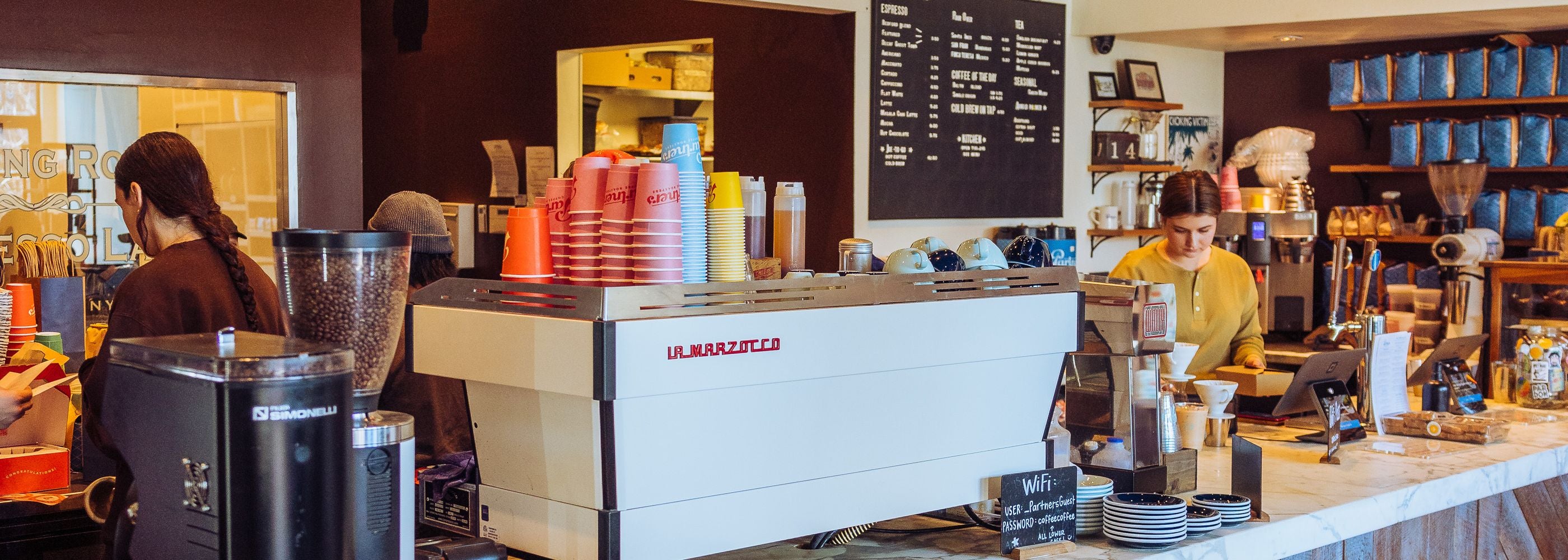 Baristas preparing espresso behind the counter at Partners Coffee Williamsburg cafe with patrons and warm interior