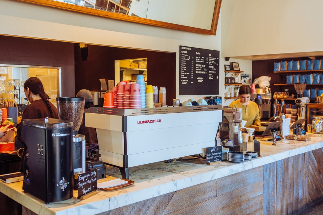 Barista preparing espresso behind a marble-top counter with a white La Marzocco machine and stacks of red takeaway cups in a bright cafe