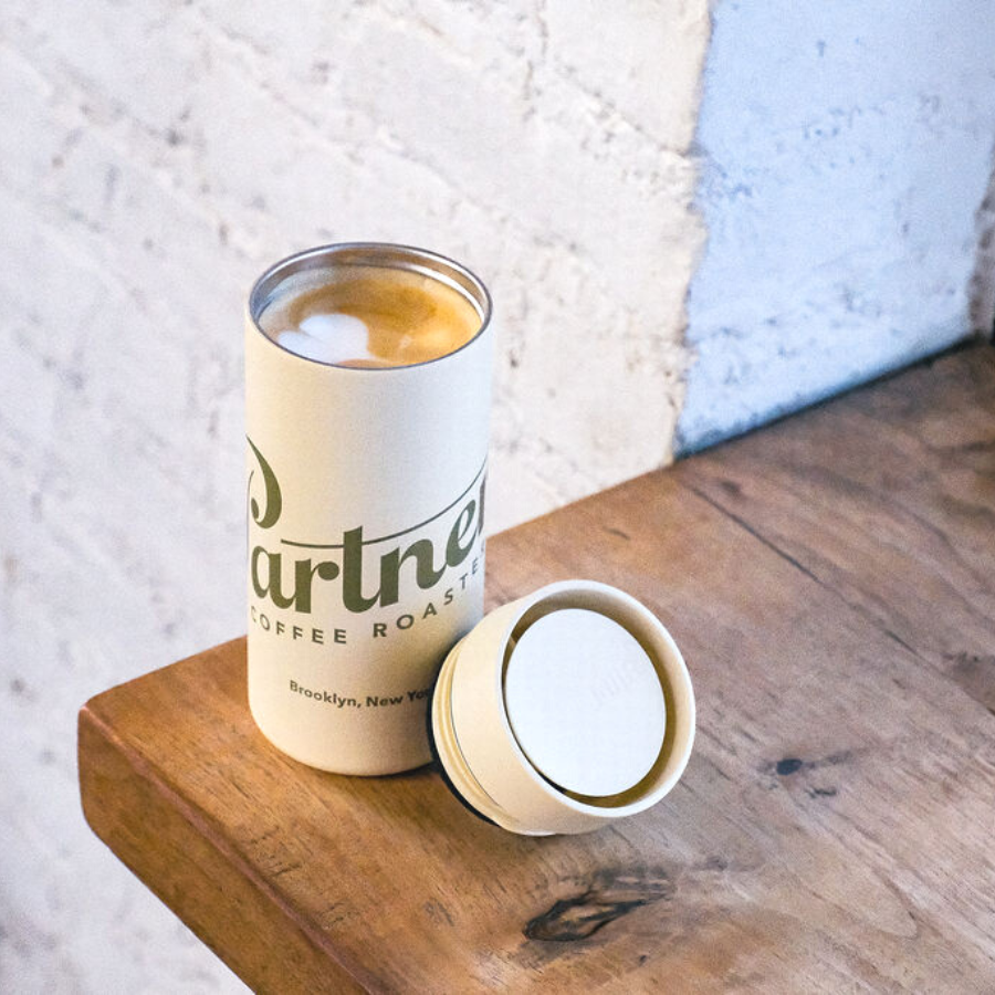 Three-quarter view of Partners x MiiR 360 Traveler mug in Sandstone with lid off showing latte foam, on a wooden table by a brick wall